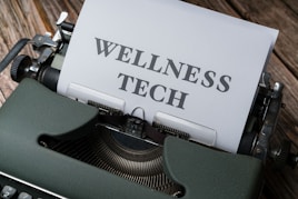 A typewriter with a sheet of paper in the carriage displaying the text 'WELLNESS TECH'. The typewriter is situated on a wooden surface, contributing to a vintage look.