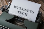 A typewriter with a sheet of paper in the carriage displaying the text 'WELLNESS TECH'. The typewriter is situated on a wooden surface, contributing to a vintage look.