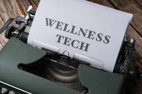 A typewriter with a sheet of paper in the carriage displaying the text 'WELLNESS TECH'. The typewriter is situated on a wooden surface, contributing to a vintage look.