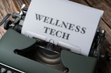 A typewriter with a sheet of paper in the carriage displaying the text 'WELLNESS TECH'. The typewriter is situated on a wooden surface, contributing to a vintage look.