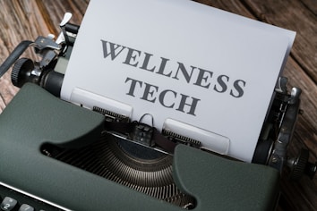 A typewriter with a sheet of paper in the carriage displaying the text 'WELLNESS TECH'. The typewriter is situated on a wooden surface, contributing to a vintage look.
