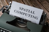 A vintage typewriter is positioned on a wooden surface with a piece of paper in the typewriter carriage displaying the text 'SPATIAL COMPUTING.' The typewriter has a dark green casing and is slightly scuffed, with metallic parts visible. The wooden surface has a rustic appearance, possibly indicating an aged or retro setting.