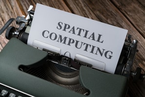 A vintage typewriter is positioned on a wooden surface with a piece of paper in the typewriter carriage displaying the text 'SPATIAL COMPUTING.' The typewriter has a dark green casing and is slightly scuffed, with metallic parts visible. The wooden surface has a rustic appearance, possibly indicating an aged or retro setting.