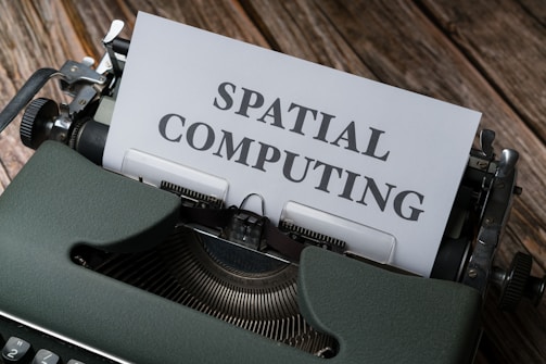 A vintage typewriter is positioned on a wooden surface with a piece of paper in the typewriter carriage displaying the text 'SPATIAL COMPUTING.' The typewriter has a dark green casing and is slightly scuffed, with metallic parts visible. The wooden surface has a rustic appearance, possibly indicating an aged or retro setting.