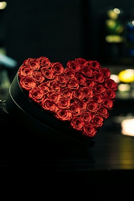 A heart-shaped arrangement of vibrant red roses placed in a box against a dark background. Soft lighting accentuates the petals' texture and the rich color of the roses.