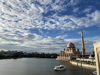 An ornate mosque with a large dome and tall minaret is situated by a calm body of water under a partly cloudy sky. A boat floats on the water, and a city skyline with various buildings is visible in the distance.