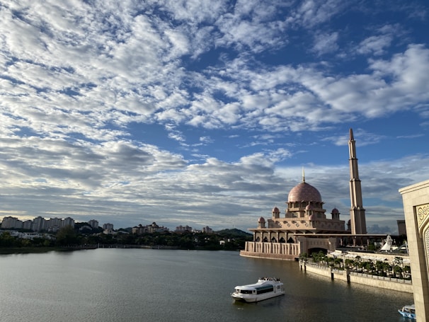 An ornate mosque with a large dome and tall minaret is situated by a calm body of water under a partly cloudy sky. A boat floats on the water, and a city skyline with various buildings is visible in the distance.