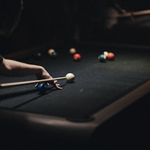 A person is playing pool on a dark billiard table, focusing intently on hitting the white cue ball with a pool stick. Several colorful balls are scattered on the table, and the scene is dimly lit, highlighting the concentration in the game.