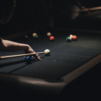 A person is playing pool on a dark billiard table, focusing intently on hitting the white cue ball with a pool stick. Several colorful balls are scattered on the table, and the scene is dimly lit, highlighting the concentration in the game.