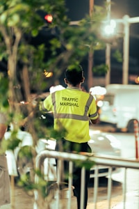 A traffic marshal is standing on the street wearing a bright yellow safety vest with reflective stripes, directing vehicles. The background shows an urban setting with blurred lights and a few cars, and the foreground includes some green foliage.