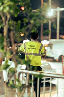 A traffic marshal is standing on the street wearing a bright yellow safety vest with reflective stripes, directing vehicles. The background shows an urban setting with blurred lights and a few cars, and the foreground includes some green foliage.