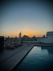 A serene rooftop pool area at sunset with lounge chairs lined up along the edge. In the background, iconic architectural structures are silhouetted against a vibrant orange and blue sky. An airplane trail is visible in the sky above.