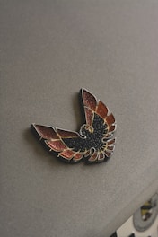 A metallic badge featuring a stylized bird with wings spread, displaying shades of black, red, and gold. The badge is laying flat on a textured grey surface, and a partial glimpse of another emblem or seal is visible at the bottom right corner.