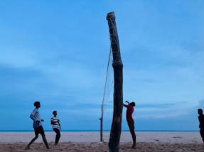 A group of people playing beach volleyball with a net set up on the sandy beach. The sky is mostly clear with a soft blue hue scattered across the horizon. The players are actively engaged in the game, casting shadows on the sand as the sun sets.