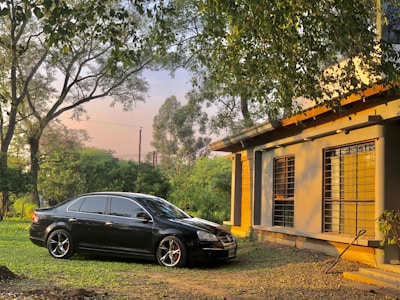 A black car is parked on a grass lawn beside a house. The house has large windows with metal bars, and the exterior is painted in a light yellow tone. Surrounding the car and house are tall trees with dense green foliage, and the sky is visible through the leaves, suggesting it is late afternoon or early evening.