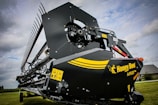 A close-up view of an agricultural harvesting machine part with a large blade and gears. The machine has a black and yellow color scheme with a logo reading 'Honey Bee AirFLEX'. Warning labels are visible on the side of the machinery. The background includes a grassy field and a cloudy sky.