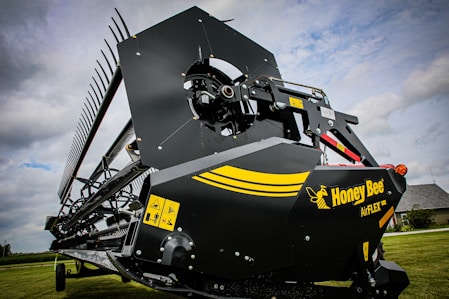 A close-up view of an agricultural harvesting machine part with a large blade and gears. The machine has a black and yellow color scheme with a logo reading 'Honey Bee AirFLEX'. Warning labels are visible on the side of the machinery. The background includes a grassy field and a cloudy sky.