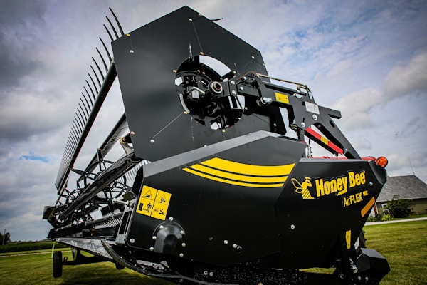 A close-up view of an agricultural harvesting machine part with a large blade and gears. The machine has a black and yellow color scheme with a logo reading 'Honey Bee AirFLEX'. Warning labels are visible on the side of the machinery. The background includes a grassy field and a cloudy sky.