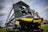 A close-up view of an agricultural harvesting machine part with a large blade and gears. The machine has a black and yellow color scheme with a logo reading 'Honey Bee AirFLEX'. Warning labels are visible on the side of the machinery. The background includes a grassy field and a cloudy sky.