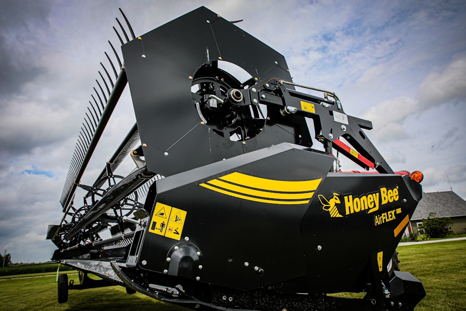 A close-up view of an agricultural harvesting machine part with a large blade and gears. The machine has a black and yellow color scheme with a logo reading 'Honey Bee AirFLEX'. Warning labels are visible on the side of the machinery. The background includes a grassy field and a cloudy sky.