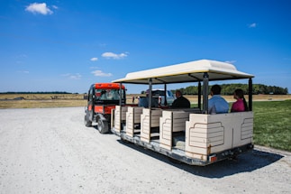A small open-air tram is being pulled by a red utility vehicle on a dirt path. The tram is carrying passengers who are looking forward, with a scenic background of a wide-open field under a clear blue sky. Sparse clouds are spread out in the sky, and the landscape includes patches of grass and a treeline in the distance.