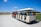 A small open-air tram is being pulled by a red utility vehicle on a dirt path. The tram is carrying passengers who are looking forward, with a scenic background of a wide-open field under a clear blue sky. Sparse clouds are spread out in the sky, and the landscape includes patches of grass and a treeline in the distance.
