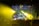A nighttime outdoor concert with a large crowd gathered in front of a stage. The stage is illuminated with vibrant yellow and green lights. A DJ booth is positioned at the center with multiple performers visible, and the audience is holding flags.