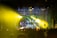 A nighttime outdoor concert with a large crowd gathered in front of a stage. The stage is illuminated with vibrant yellow and green lights. A DJ booth is positioned at the center with multiple performers visible, and the audience is holding flags.