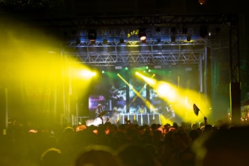 A nighttime outdoor concert with a large crowd gathered in front of a stage. The stage is illuminated with vibrant yellow and green lights. A DJ booth is positioned at the center with multiple performers visible, and the audience is holding flags.