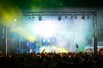 A vibrant outdoor concert scene with a large crowd gathered in front of a stage illuminated with bright lights and colored beams. The atmosphere is lively and energetic, with people raising their hands in the air. Smoke and laser lights create a dynamic and festive environment.
