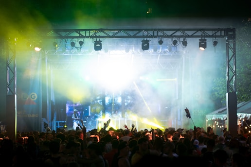 A vibrant outdoor concert scene with a large crowd gathered in front of a stage illuminated with bright lights and colored beams. The atmosphere is lively and energetic, with people raising their hands in the air. Smoke and laser lights create a dynamic and festive environment.