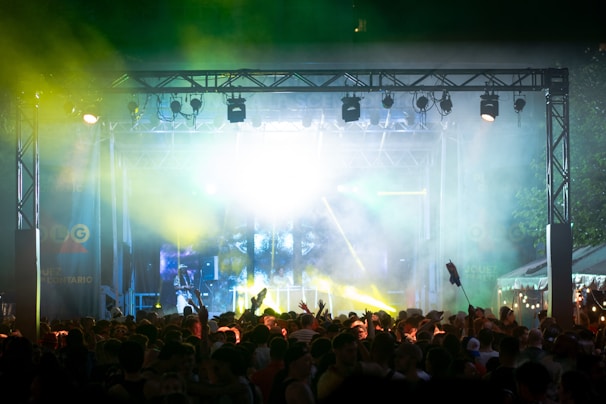 A vibrant outdoor concert scene with a large crowd gathered in front of a stage illuminated with bright lights and colored beams. The atmosphere is lively and energetic, with people raising their hands in the air. Smoke and laser lights create a dynamic and festive environment.