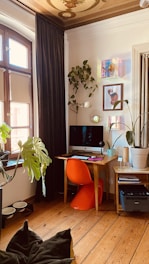 A cozy workspace features a wooden desk with a computer on it, accompanied by an orange chair. The area is surrounded by decorative plants, and artworks are hung on the white walls. Large windows with brown frames and heavy curtains let in natural light, illuminating the wooden floor. A brightly patterned ceiling adds elegance to the room.