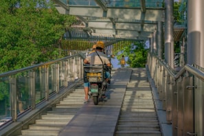 A delivery person on a motorbike is riding up a ramp with glass railings on both sides. The scene is set in a sunny outdoor environment with green trees visible in the background. A pedestrian is walking under a covered walkway ahead.
