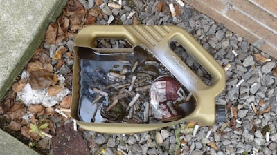 A discarded plastic container is filled with dirty water and cigarette butts. It is lying on the ground amidst stones, dry leaves, and other debris near a brick wall.