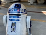 A metallic robot with a white and blue cylindrical body and dome-shaped head. The robot has various panels and components on its body, along with a lens-like feature on the upper part. The background appears to be an industrial or mechanical setting.