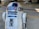 A metallic robot with a white and blue cylindrical body and dome-shaped head. The robot has various panels and components on its body, along with a lens-like feature on the upper part. The background appears to be an industrial or mechanical setting.