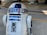A metallic robot with a white and blue cylindrical body and dome-shaped head. The robot has various panels and components on its body, along with a lens-like feature on the upper part. The background appears to be an industrial or mechanical setting.