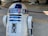 A metallic robot with a white and blue cylindrical body and dome-shaped head. The robot has various panels and components on its body, along with a lens-like feature on the upper part. The background appears to be an industrial or mechanical setting.