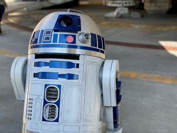 A metallic robot with a white and blue cylindrical body and dome-shaped head. The robot has various panels and components on its body, along with a lens-like feature on the upper part. The background appears to be an industrial or mechanical setting.