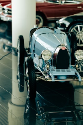 A vintage-style toy car with a classic design is positioned on a reflective tiled floor. The blue car features a distinctive front grille, round headlights, and thin tires. In the background, parts of two other vehicles, including a red one, can be seen, suggesting a showroom setting.