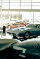 Several classic sports cars are displayed in what appears to be a showroom with large windows. The cars have sleek, vintage designs, and the lighting creates a reflective surface on the polished floor.