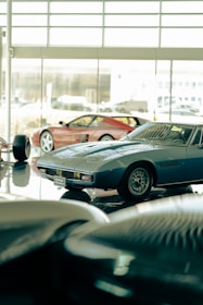 Several classic sports cars are displayed in what appears to be a showroom with large windows. The cars have sleek, vintage designs, and the lighting creates a reflective surface on the polished floor.