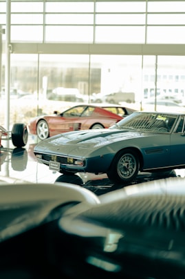 Several classic sports cars are displayed in what appears to be a showroom with large windows. The cars have sleek, vintage designs, and the lighting creates a reflective surface on the polished floor.