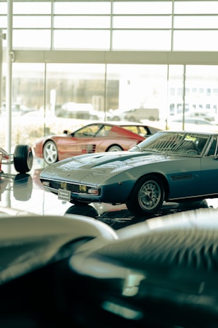 Several classic sports cars are displayed in what appears to be a showroom with large windows. The cars have sleek, vintage designs, and the lighting creates a reflective surface on the polished floor.