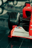 A close-up view of the front section of a red Formula 1 race car featuring a large black tire, branding and sponsorship logos such as FedEx, Bridgestone, and Shell on the white front wing. The car is displayed on a glossy, reflective surface.