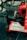 A close-up view of the front section of a red Formula 1 race car featuring a large black tire, branding and sponsorship logos such as FedEx, Bridgestone, and Shell on the white front wing. The car is displayed on a glossy, reflective surface.