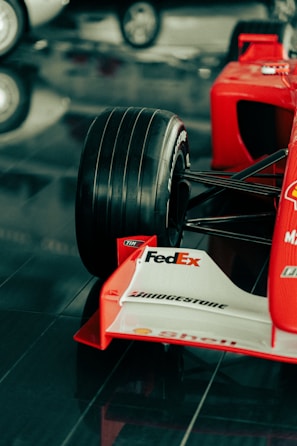 A close-up view of the front section of a red Formula 1 race car featuring a large black tire, branding and sponsorship logos such as FedEx, Bridgestone, and Shell on the white front wing. The car is displayed on a glossy, reflective surface.
