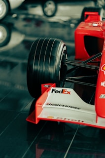 A close-up view of the front section of a red Formula 1 race car featuring a large black tire, branding and sponsorship logos such as FedEx, Bridgestone, and Shell on the white front wing. The car is displayed on a glossy, reflective surface.