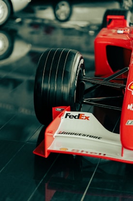 A close-up view of the front section of a red Formula 1 race car featuring a large black tire, branding and sponsorship logos such as FedEx, Bridgestone, and Shell on the white front wing. The car is displayed on a glossy, reflective surface.